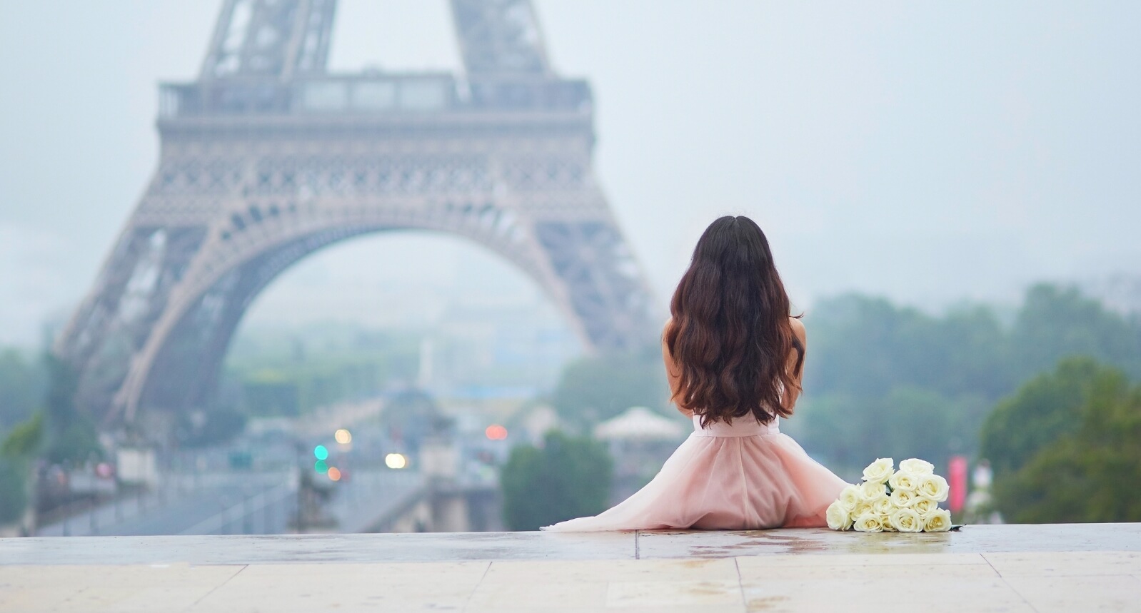Woman in a pink dress sitting with a bouquet of white roses overlooking the Eiffel Tower, evoking the romantic aura of French inspired candles.