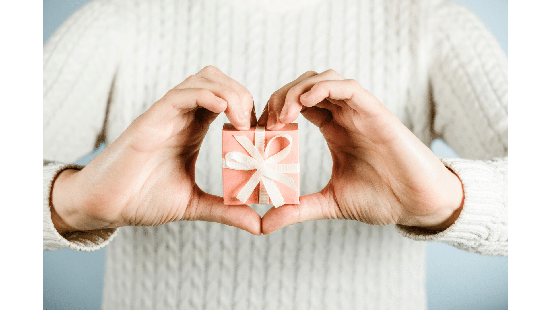 Woman holding a small gift box with a white ribbon, forming a heart shape with her hands around it highlighting unique gift ideas.